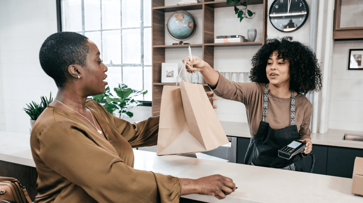 an african american employee smiling and handing a paper shopping bag over the counter to a customer